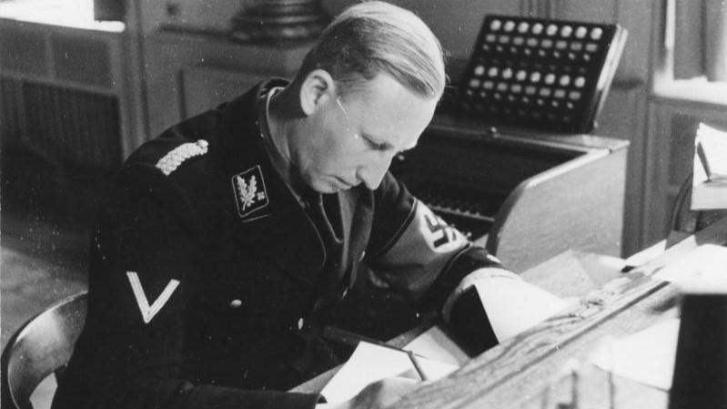 Black-and-white photo of SS officer Reinhard Heydrich seated at a desk, writing documents in his Munich office in 1934.